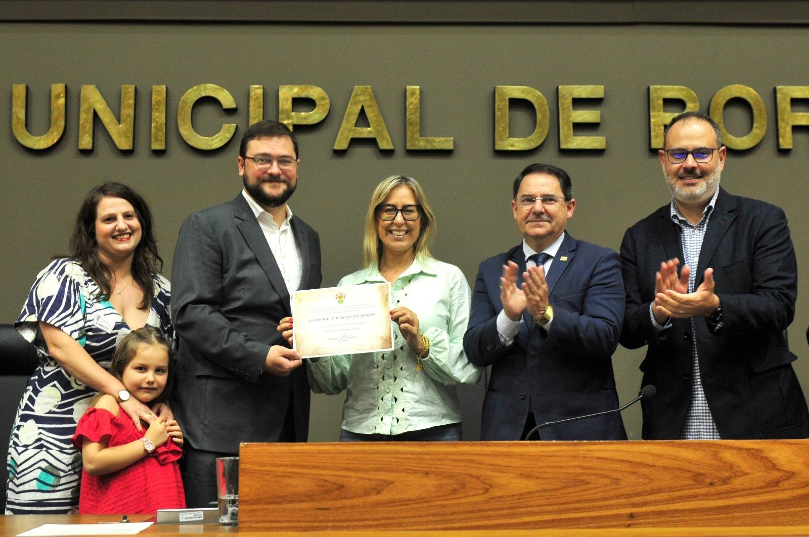 O diploma foi entregue ao jornalista Guilherme Buamhardt (E) pela vereadora Fernanda Barth (D), ao lado do presidente da c&acirc;mara Hamilton Sossmeier e o vice-prefeito Ricardo Gomes. Na foto, a esquerda a esposa de Baumhardt, Janine e a filha, Elisa.