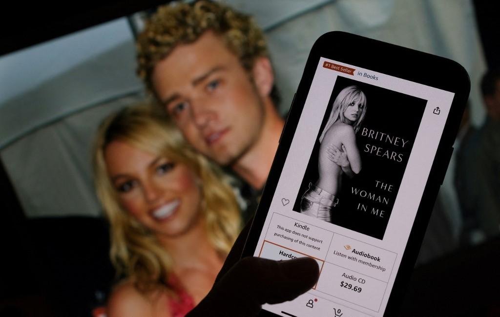 Capa do livro "The Woman in Me"  de uma loja online na frente de uma foto de Britney Spears e Justin Timberlake no American Music Awards de 9 de janeiro de 2002
