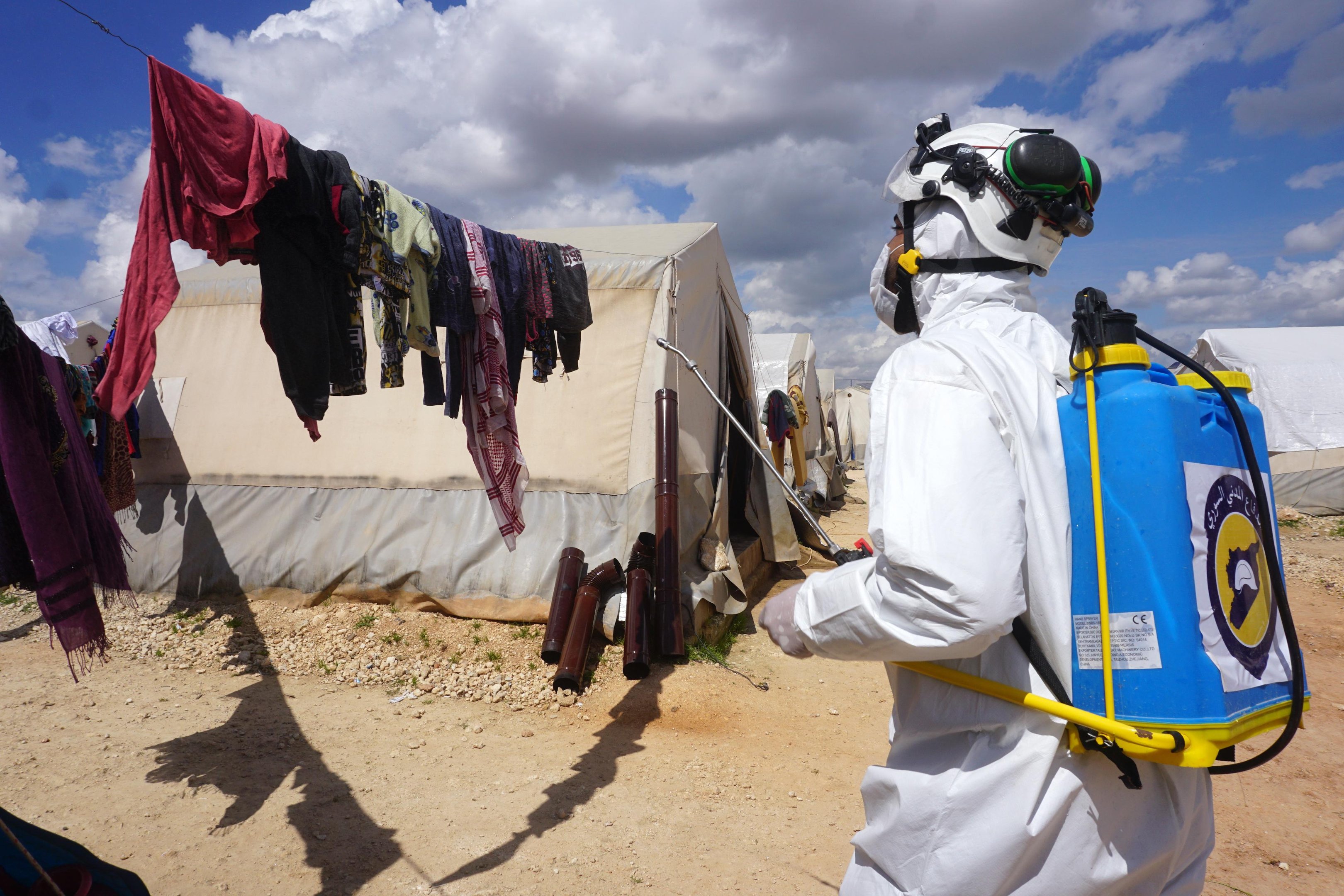 Um membro da defesa civil s&iacute;ria pulveriza desinfetante nas roupas penduradas do lado de fora de uma tenda em um campo para pessoas deslocadas perto da cidade de Maaret Misrin