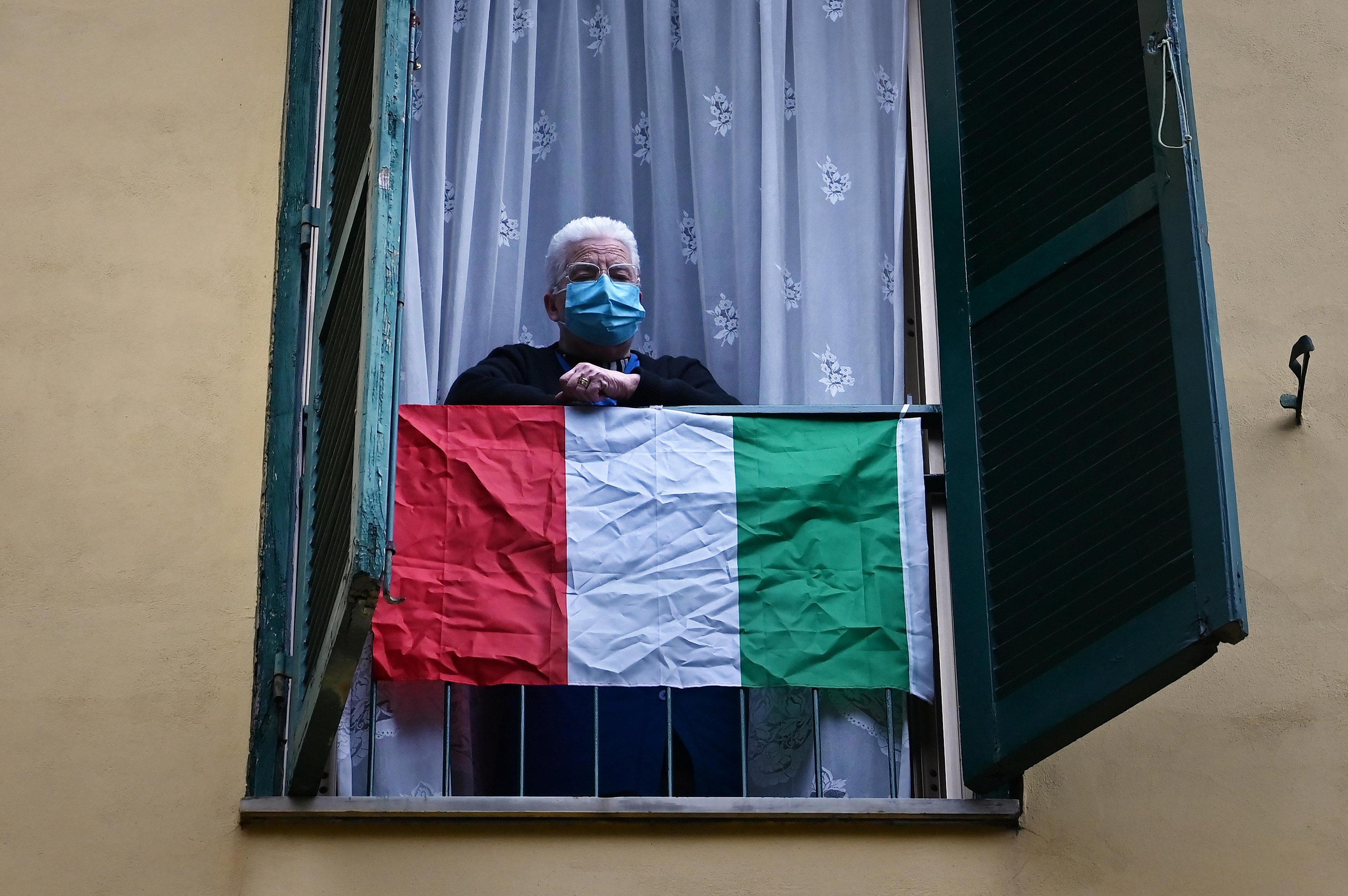 Um idoso usando uma m&aacute;scara facial fica atr&aacute;s de uma bandeira nacional italiana enquanto artistas se apresentam no p&aacute;tio de um pr&eacute;dio de apartamentos popular para o show Sotto lo Stesso Cielo tour, nos sub&uacute;rbios de San Basilio, em Roma