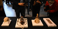 Entre os objetos estão um vaso decorado com três cabeças, uma boneca em trajes tradicionais, vários acessórios de cabelo e lanças